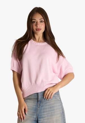 Giovane donna con lunghi capelli castani che indossa un maglione rosa chiaro a maniche corte e jeans blu chiaro, posando davanti a uno sfondo bianco.