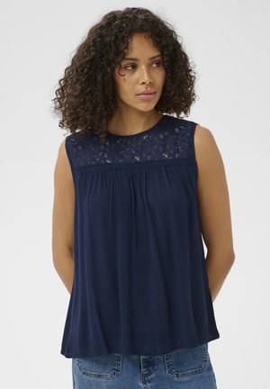 Femme aux cheveux bouclés et foncés portant un haut bleu marine sans manches avec un empiècement en dentelle et un jean en denim bleu, tournée légèrement vers la gauche.