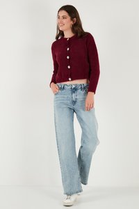 Giovane donna sorridente, indossa un cardigan bordeaux morbido con bottoni, jeans a gamba larga azzurri e sneakers bianche, in piedi con una mano in tasca.