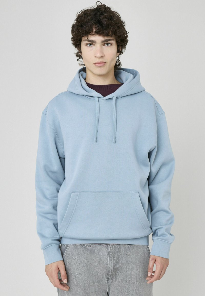 Haut à capuche bleu clair en tissu doux avec une poche avant, un cordon de serrage et des poignets côtelés. Porté par-dessus une chemise bordeaux foncé.