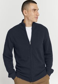 Marinblå zip-up tröja med texturerade axelpaneler, ribbade ärmslut och nederkant. Bärs över en vit skjorta och kompletterar en neutral bakgrund.