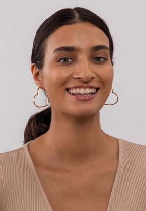 Gouden hartvormige hoop oorbellen, open ontwerp, lichtgewicht en met een gestructureerd detail, gedragen door een model met een neutrale blouse.