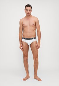 Chiloți scurți albi cu un tiv negru, având logo-ul "justcavalli." Modelul stă desculț, cu fața spre cameră, evidențiind un design ajustat.