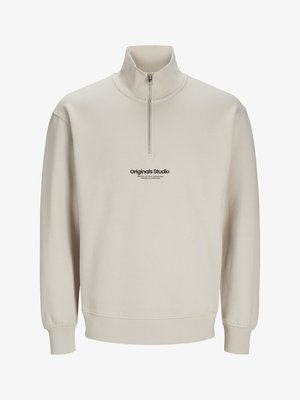 Beżowa rozpinana bluza z wysokim kołnierzem, ściągaczami przy mankietach oraz nadrukiem logo "Originals Studio" z przodu. Miękka tekstura materiału.