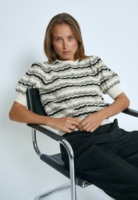 Texturiertes Pulli in Creme mit schwarzen Zickzackstreifen, ausgestattet mit kurzen, puffartigen Ärmeln und einem gerippten Halsausschnitt, kombiniert mit schwarzen Hosen.