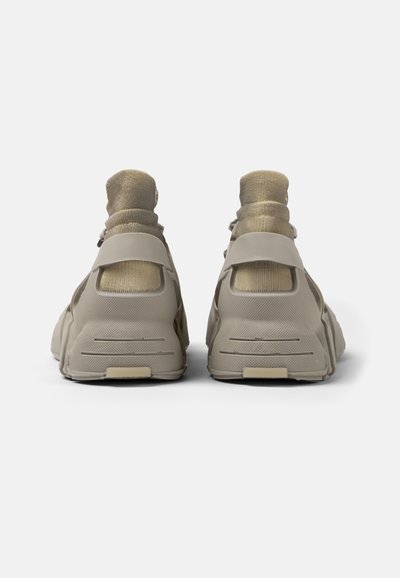 Scarpe sportive beige con una superficie testurizzata, tomaia simile a un calzino e cinturini regolabili sul tallone. Suole piatte con lievi scanalature per una maggiore aderenza.