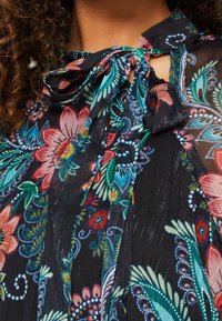 Närbild av en blommönstrad genomskinlig blus med knuten rosettkrage i nyanser av rött, grönt, blått och svart på en person med lockigt hår.