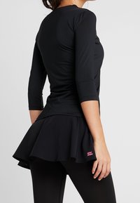 Svart träningslinne med trekvartslånga ärmar och ett utsvängt nederkant, med en texturerad yta och en rosa logotypdetalj.