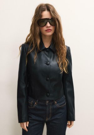 Kvinde med langt, bølget hår iført mørke, store solbriller, sort læderjakke med knapper og mørke denimjeans foran en ensfarvet baggrund.