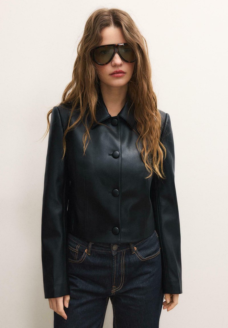 Frau mit langen, welligen Haaren, die dunkle übergroße Sonnenbrillen, eine schwarze Lederjacke mit Knöpfen und dunkle Jeans trägt, vor einfarbigem Hintergrund.