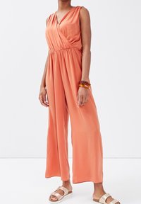 Combinaison de couleur pêche avec un design à l'avant croisé, sans manches, et une taille élastique. Accompagnée de sandales beige et d'un bracelet ambre.