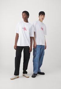 Dva jedinci oblečení v bílých bavlněných tričkách s červeným logem Calvin Klein. Jeden v černých kalhotách, druhý ve světle modrých denimových cargo kalhotách.