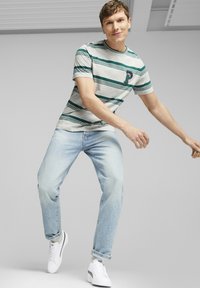 T-shirt cinza e verde às riscas com gola redonda e um logótipo no peito, combinada com jeans azul claro e sapatilhas brancas, mostrada em movimento.