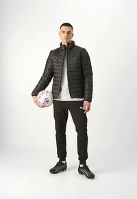 Melnā polsterētā jaka, pelēka T-krekls, melnas sporta bikses, rokās tur daudzkrāsainu futbola bumbu, valkā melnus futbola zābakus.