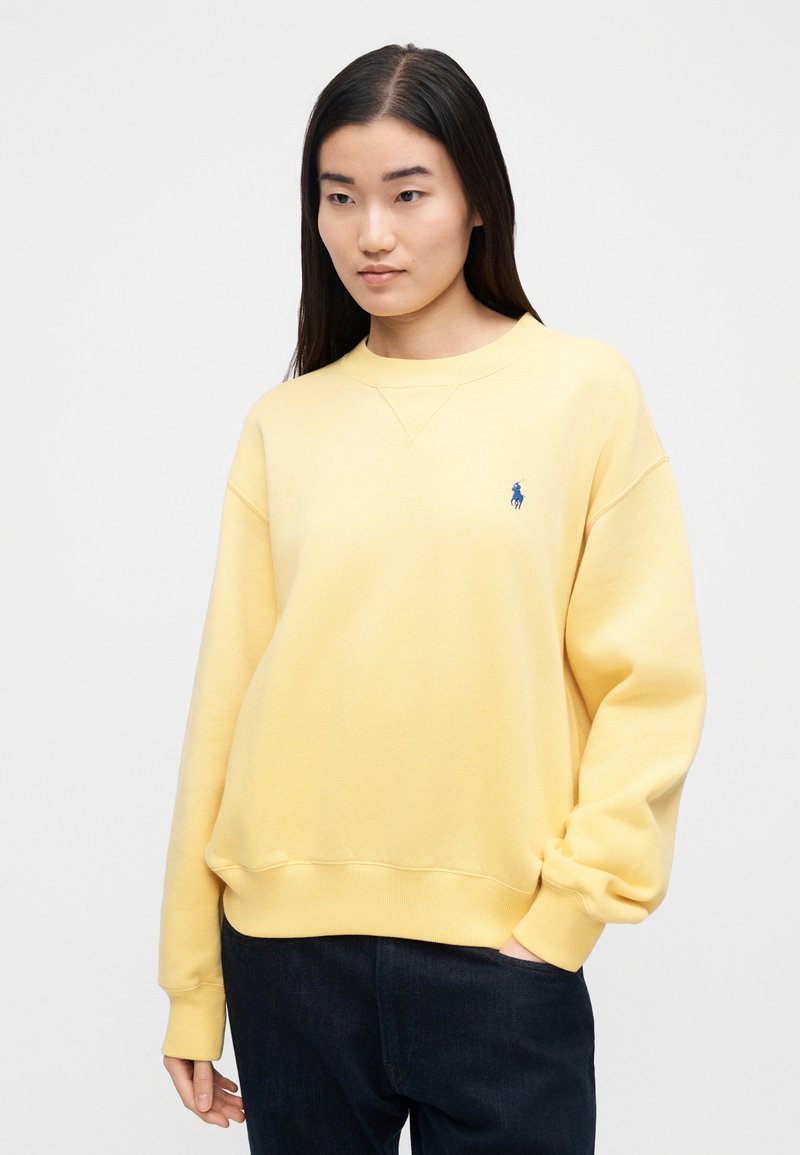 Sweat-shirt jaune avec un col rond et des poignets côtelés, comportant un petit logo bleu sur le côté gauche de la poitrine. Texture douce, coupe décontractée.