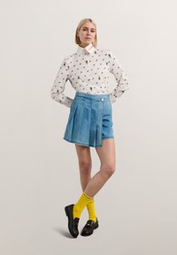 Hvit skjorte med blomstertrykk, kombinert med en lys blå, plisserte jeanskjørt og svarte mokkasiner. Gule sokker gir et fargerikt preg.