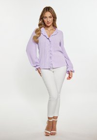 Lila fodros blúz hosszú ujjal, gombos elülső résszel, texturált anyagból, fehér slim fit farmerrel és pántos magassarkúval párosítva.