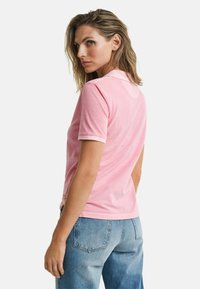 Rosa Poloshirt mit klassischem Kragen, kurzen Ärmeln und weißen Akzenten, kombiniert mit hellblauen Jeans. Glatte Textur und lässiges Design.