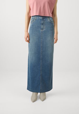 Femme portant une jupe maxi en jean bleu taille haute avec coutures, poches avant et chaussures pointues beiges, associée à un top court rose.