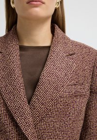 Blazer con una tela tejida texturada en púrpura y beige, con un cuello muesca y un bolsillo en el pecho, superpuesto a una blusa marrón.