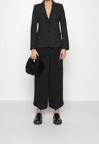 Black tailored blazer with sharp lapels, two buttons, and front pockets, paired with wide-leg trousers. Holding a round, textured black bag.