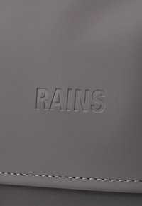 Sac gris avec logo "RAINS" en relief, comportant un matériau lisse et de fines coutures. Le design simple met l'accent sur la texture élégante et l'attrait moderne.
