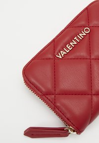 Κόκκινο δέρμα πορτοφόλι με επένδυση καπιτονέ, χρυσά αξεσουάρ και κλείσιμο με φερμουάρ. Το όνομα της μάρκας "Valentino" είναι prominently εμφανές.