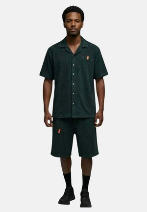 Homme debout de face portant une chemise boutonnée à manches courtes vert foncé et un short assorti longueur genoux avec des chaussettes et des chaussures noires.