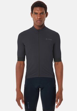 Uomo che indossa una maglia da ciclismo nera aderente e pantaloncini, in piedi di fronte con le braccia rilassate ai lati, su uno sfondo semplice.