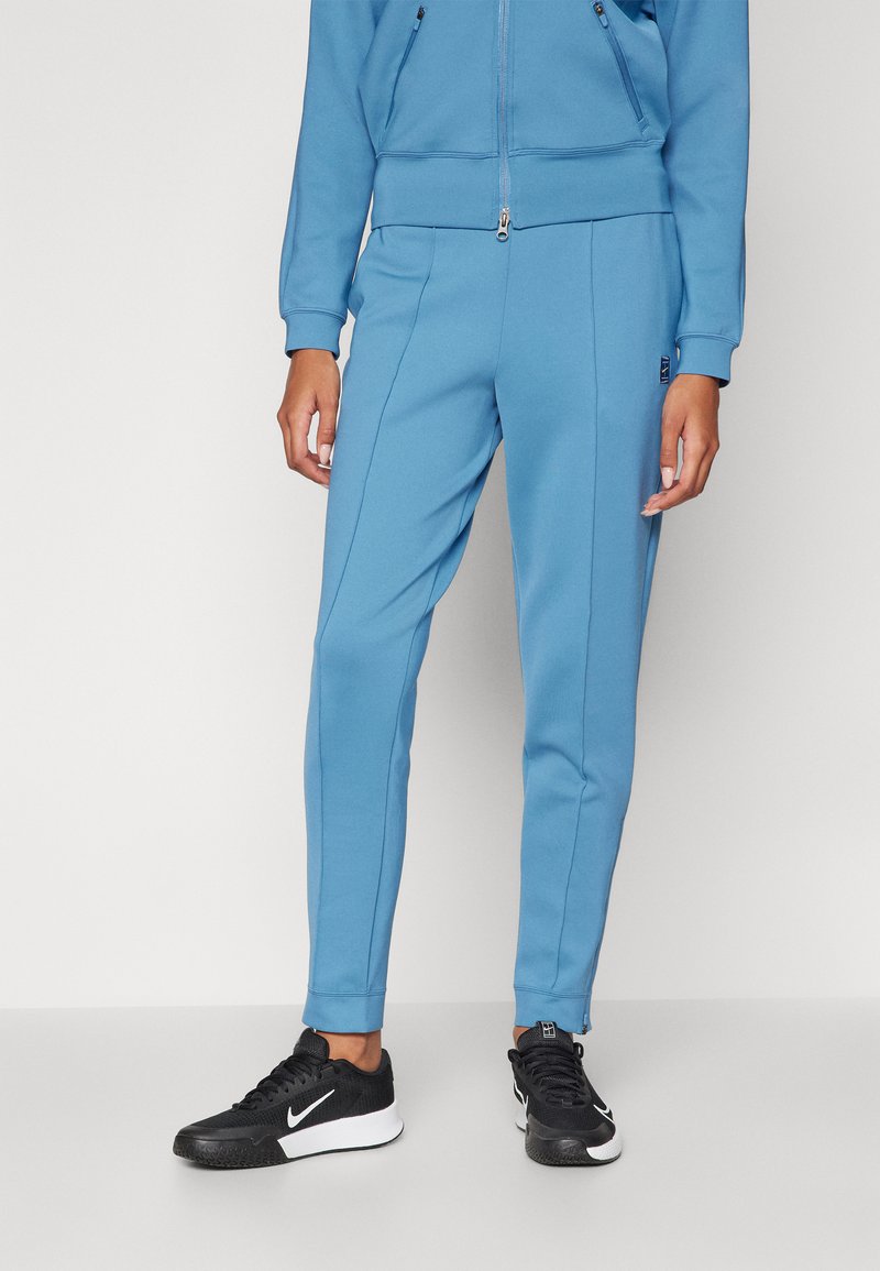 Top in tessuto tecnico azzurro chiaro con zip, abbinato a pantaloni jogger coordinati con cuciture laterali e polsini a coste. Indossato con sneakers nere.