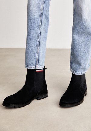 Osoba w jasnoniebieskich dżinsach i czarnych botkach za kostkę z wytrzymałymi podeszwami i elastycznymi bokami, stojąca na beżowej podłodze.