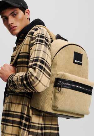 Rucsac din corduroy de culoare bej, cu formă rotundă, buzunar cu fermoar negru și etichetă cu marcă. Modelul poartă o cămașă în carouri bej și negru.