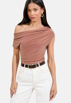 Modelka pózuje v hnědém topu bez ramínek a bílých kalhotách, doplněná náušnicemi ve tvaru kruhu a opaskem, předvádějící stylový outfit.