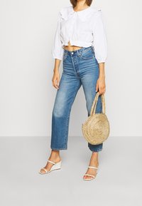 Vit kortärmad blus med volangkrage, ihop med högmidjade ljusblå jeans. Modell som håller en rund strandväska och bär vita kilklackar.