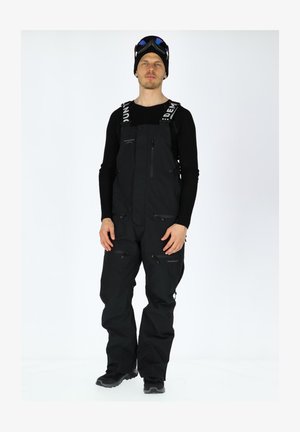 Black waterproof ski overalls with adjustable straps, multiple zippered pockets, and a loose fit. Worn with a black long-sleeve shirt and goggles.