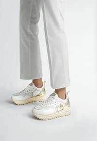 Zapatillas blancas con acentos dorados y una suela gruesa, que presentan materiales de malla y cuero, puntera redonda y seis ojales para los cordones.