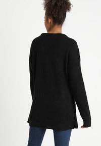 Pull en tricot côtelé noir avec des manches longues, un col rond et des fentes sur les côtés, offrant une coupe ample et un tissu texturé.