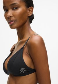 Juodas trikampis liemenėlė su plonomis reguliuojamomis petnešomis ir mažu logotipo detaliu šone. Pagaminta iš lygios medžiagos su matinio paviršiaus apdaila.