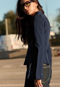 Granatowa dopasowana kurtka z guzikami i usztywnionym kołnierzem, zestawiona z ciemnymi dżinsami. Modelka nosi oversize'owe okulary z żółtymi szkłami.