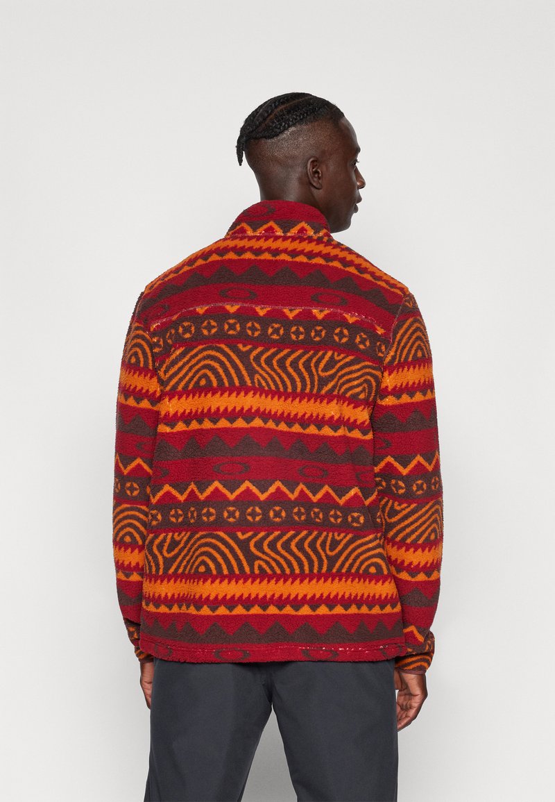 Fleece jacket featuring a vibrant red, orange, and brown pattern with geometric shapes. The design includes intricate swirls and horizontal lines.