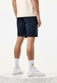 Námornícke modré šortky s klasickým strihom, nosené s bielymi ponožkami a teniskami, s akcentom usmievajúcej sa tváre na ponožke. Pozadie z mramorového podstavca.