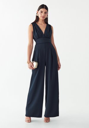 Femme portant une combinaison noire sans manches à jambes larges, tenant une petite pochette dorée, portant des boucles d'oreilles dorées et des sandales, debout sur fond blanc.