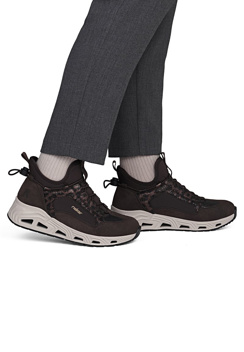 Personne portant des baskets de sport marron foncé à semelles épaisses blanches, des chaussettes claires et un pantalon gris foncé, debout sur fond blanc.