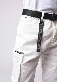 Pantalones cargo blancos con bolsillos, herrajes negros y un cinturón negro texturizado. Presenta una etiqueta con "PROJECT PARIS" en letras grises.