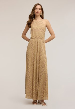 Kvinde iført en ærmeløs beige maxikjole med zigzag-mønster og halterneck samt åbne hvide hæle, står med hænderne bag ryggen.