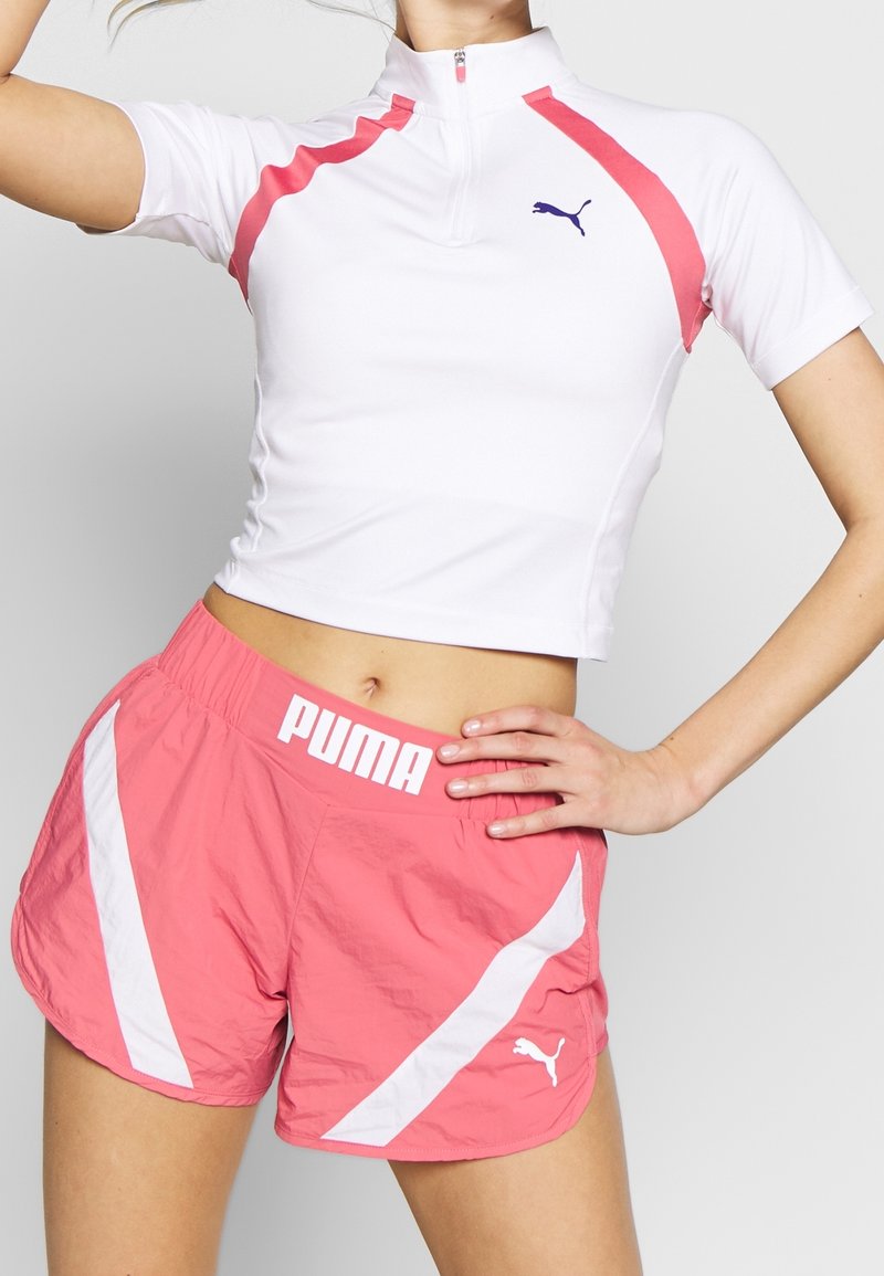 Bílý sportovní top s krátkým rukávem a růžovými akcenty, se zipovým límcem, zkombinovaný s růžovými šortkami s bílými pruhy a logem Puma na pásku.