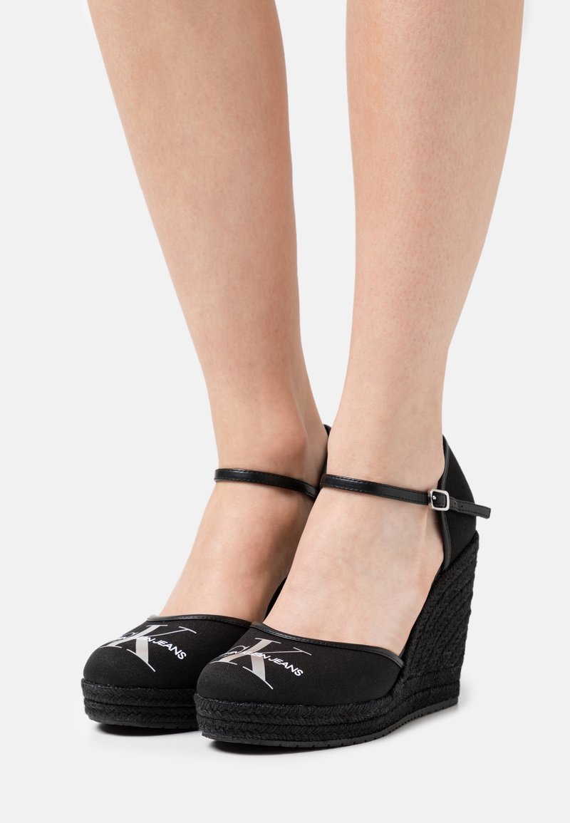 Personne portant des espadrilles compensées noires avec des brides à la cheville et un logo blanc sur le devant.