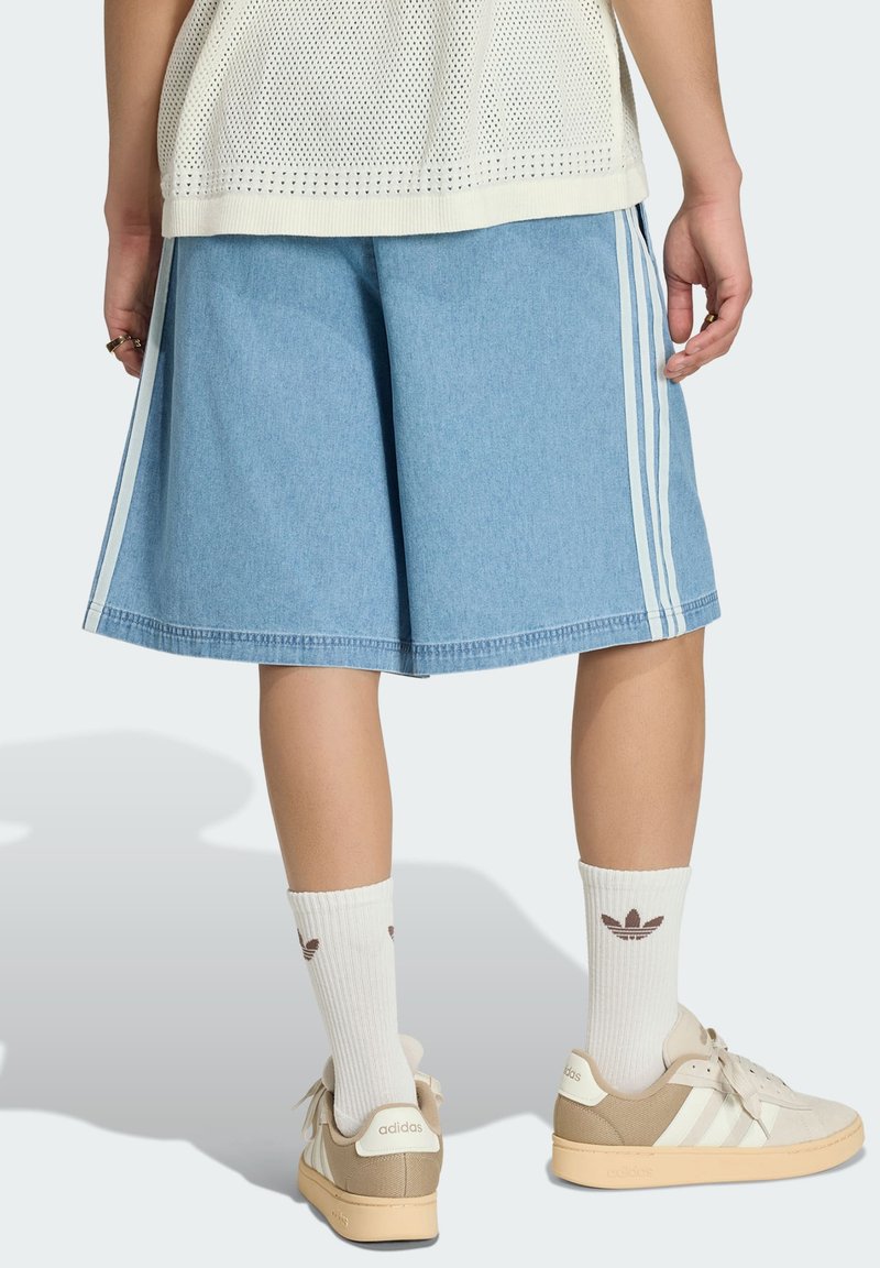 Person, die hellblaue knielange Shorts mit weißen Streifen, weiße Adidas-Socken, beige Adidas-Sneaker und ein weißes Stricktop trägt.