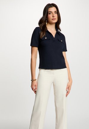 Kvinde med langt mørkt hår iført en marineblå kortærmet skjorte med krave og lommer samt hvide, vide bukser, stående mod en ensfarvet baggrund.