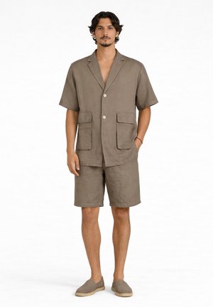 Uomo che indossa una camicia taupe a maniche corte con bottoni e due tasche, pantaloncini abbinati e scarpe slip-on, in piedi su sfondo bianco.